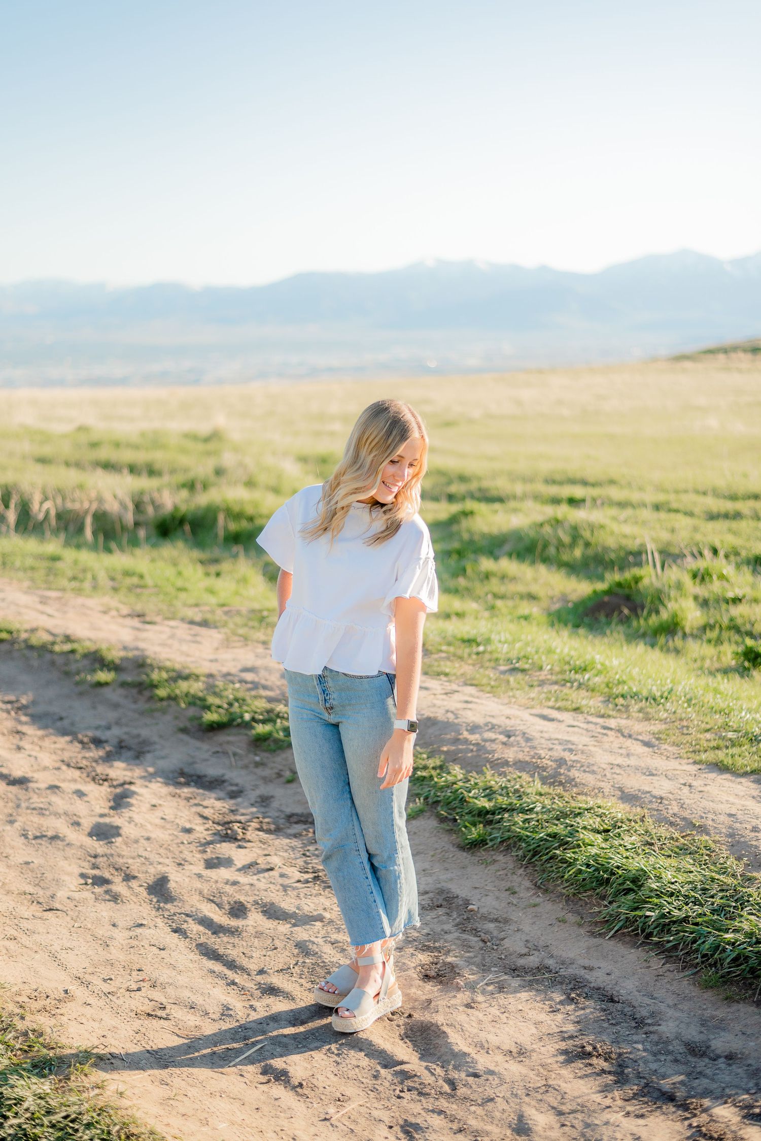 Blonde woman in white top and jeans strolling on sunny dirt path with green meadow and mountains behind. Megan Snell Photo Utah Weddings