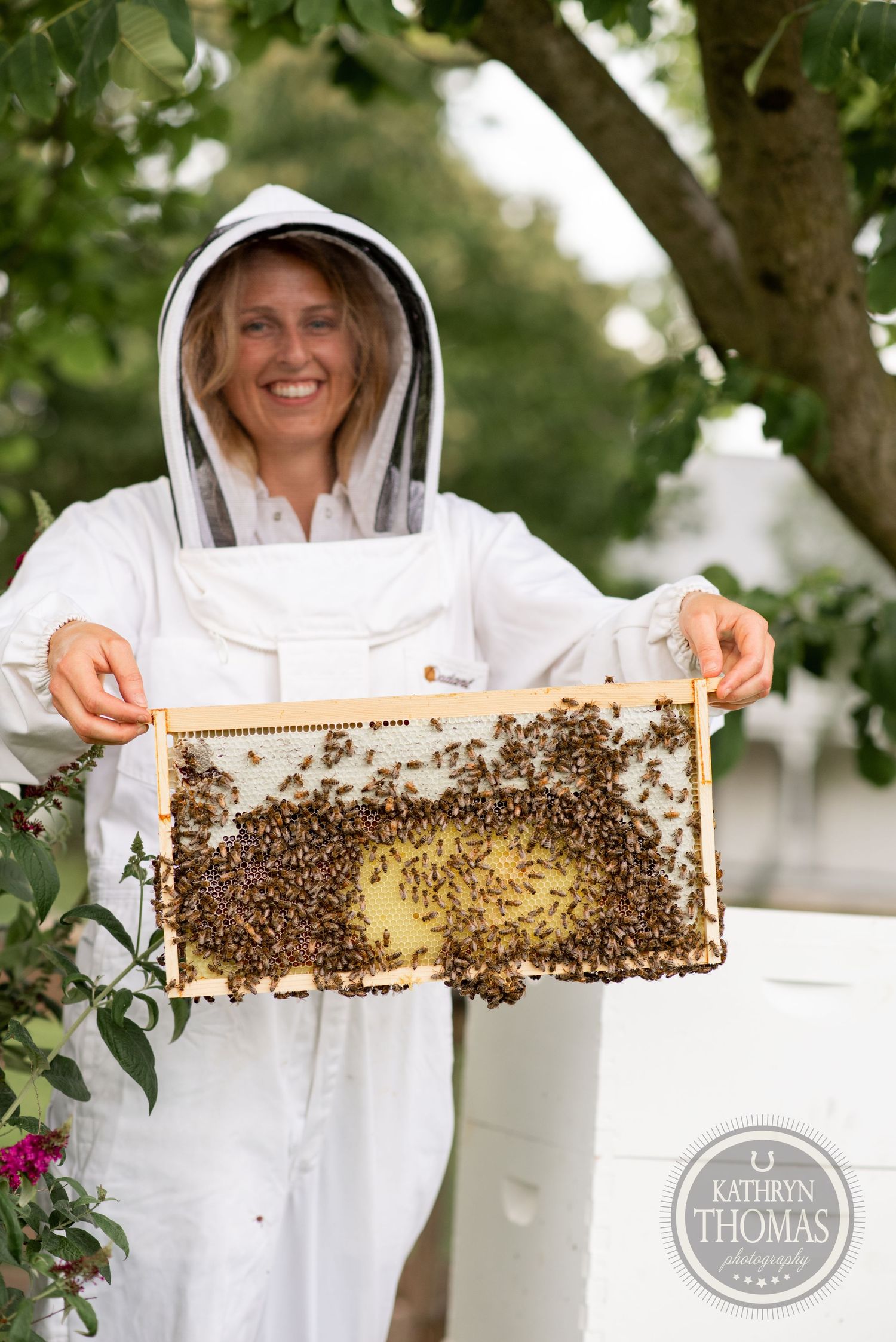 A beekeeper in protective gear proudly displays a honeycomb frame covered in busy bees.