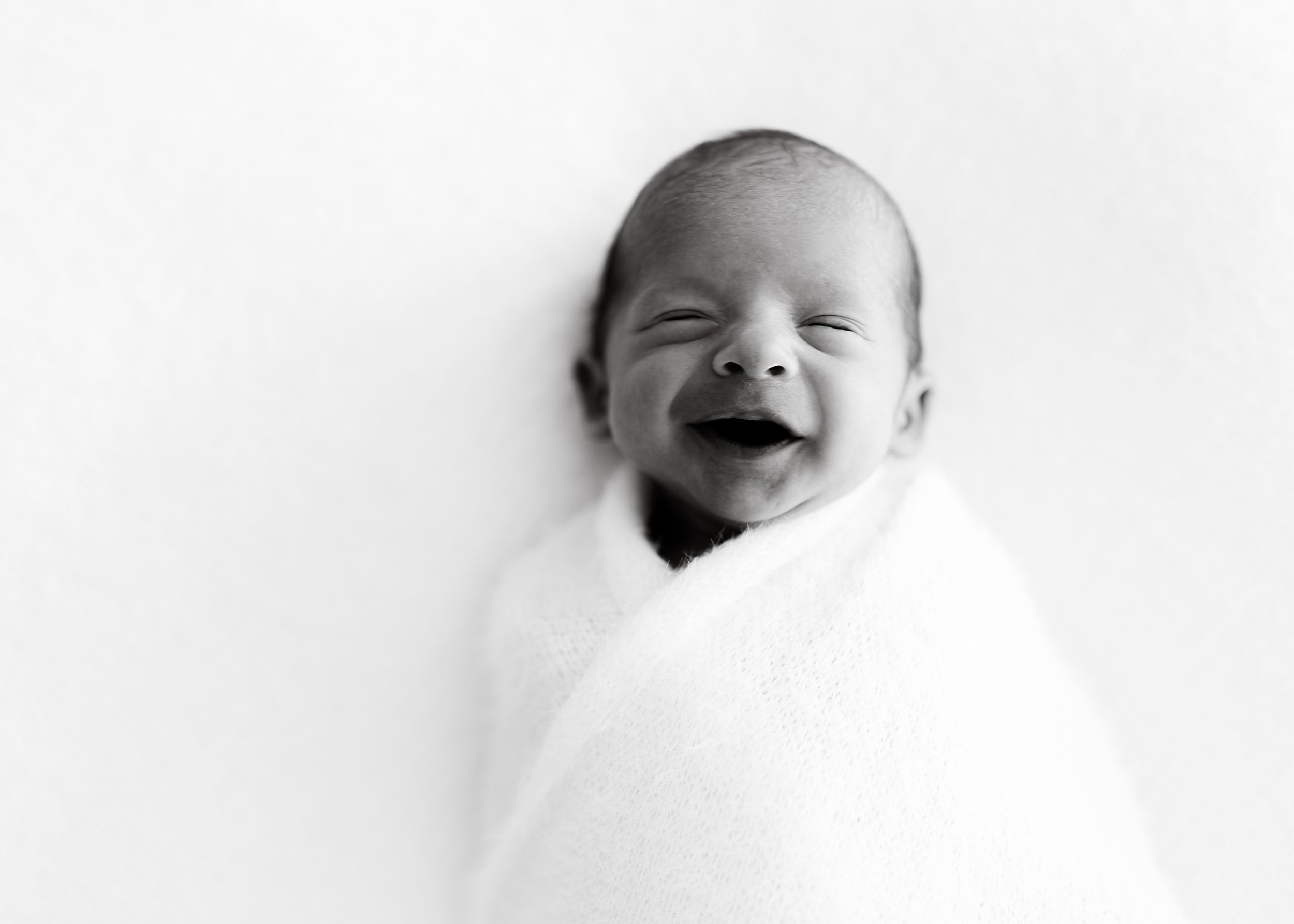 Black and white portrait of joyful newborn wrapped in white blanket.