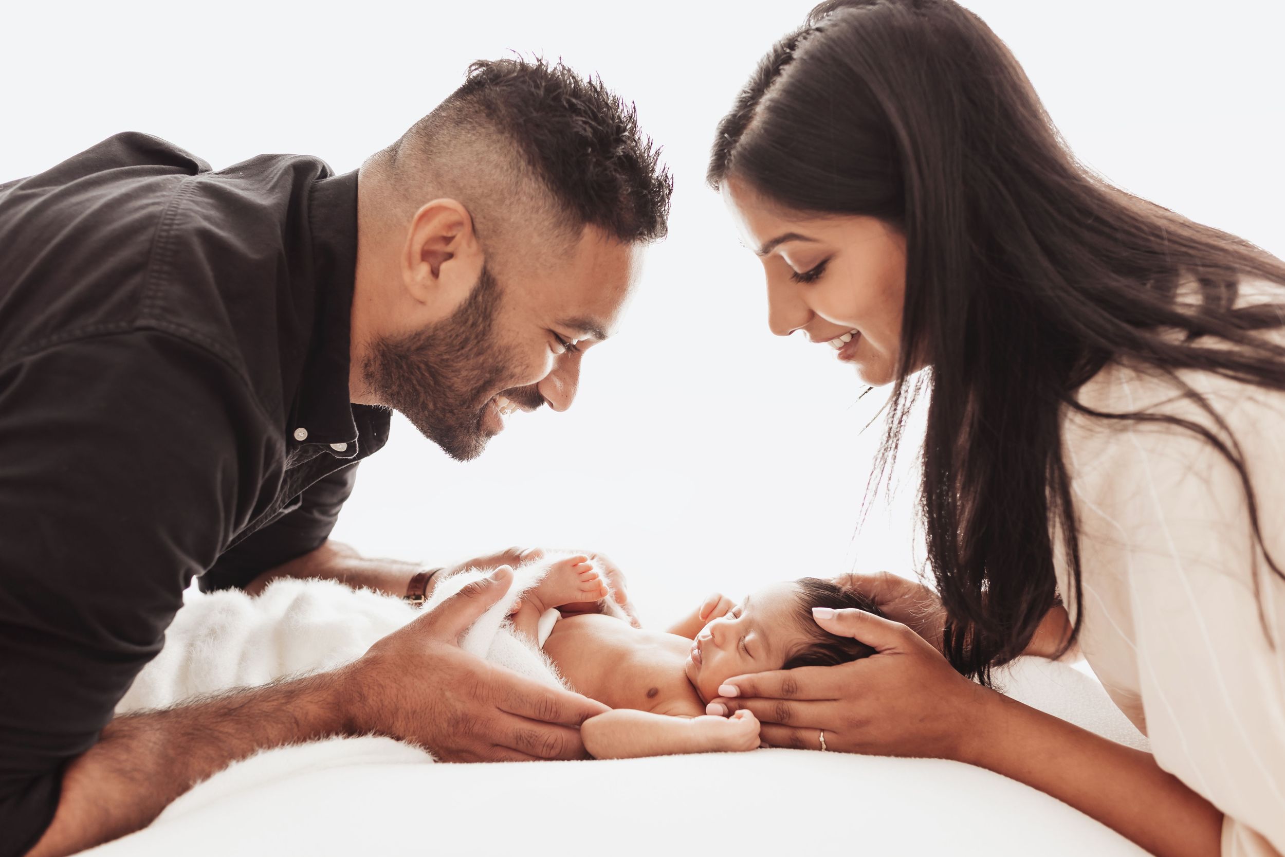 Parents lovingly gazing at their newborn baby lying between them on white background.