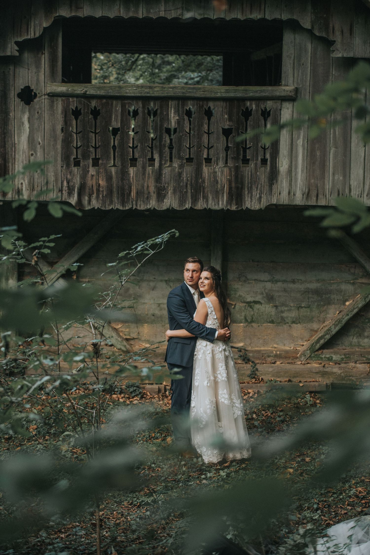Wedding couple posing beneath a rustic wooden structure in a moody forest setting.