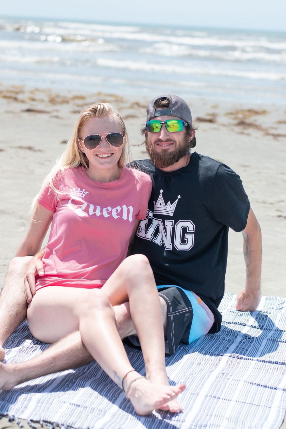 Couple in matching King and Queen shirts relaxing on blanket at beach.