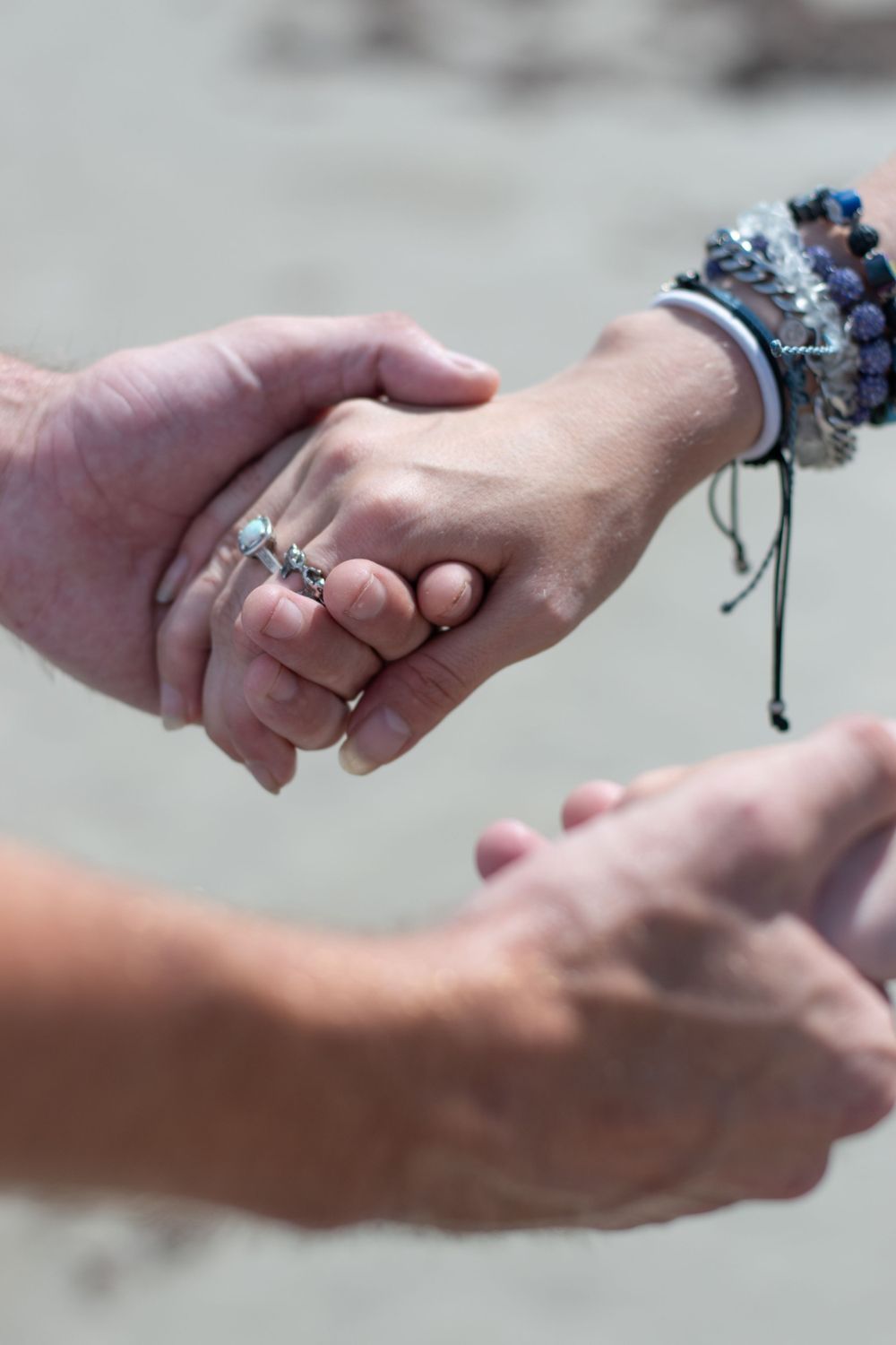 Close-up of hands wearing rings and friendship bracelets intertwined on beach.