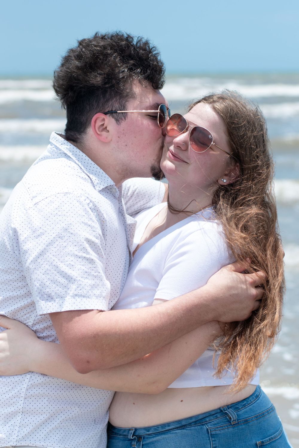 A romantic couple embraces and shares a sweet kiss on the beach with ocean waves in the background.
