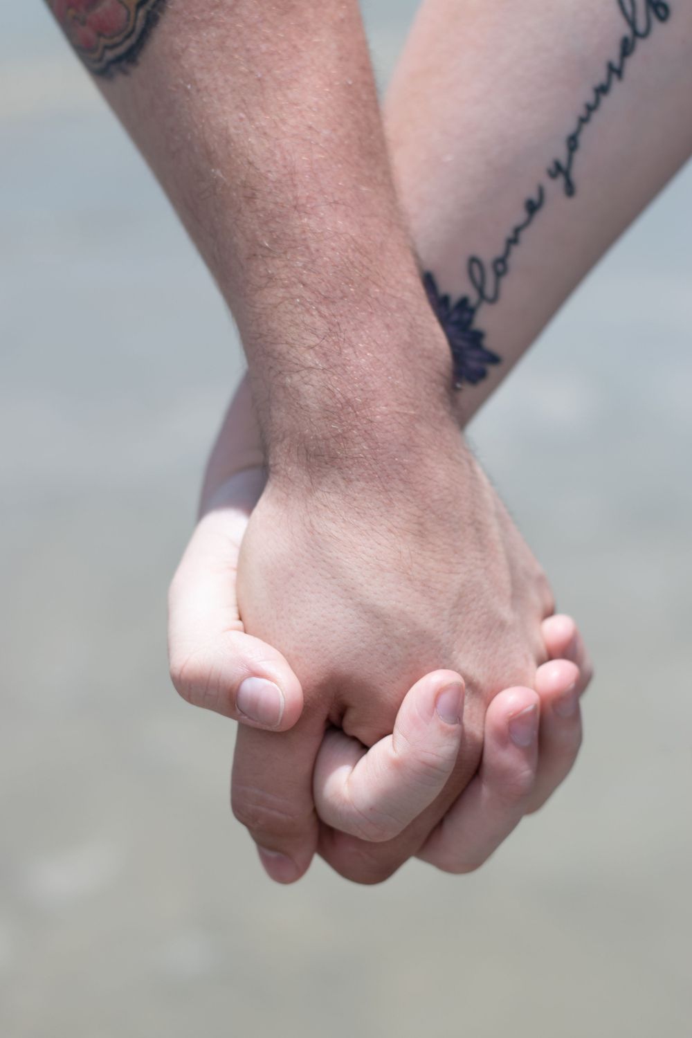 Close-up of two hands clasped together showing tattoos against a beach background.
