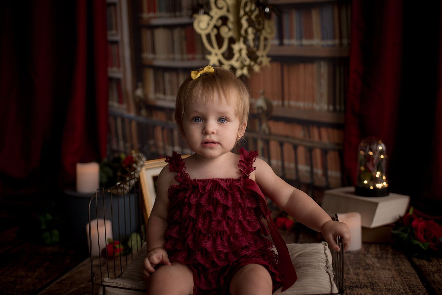 Holiday portrait of toddler in red romper sitting in library setting with golden decorations.