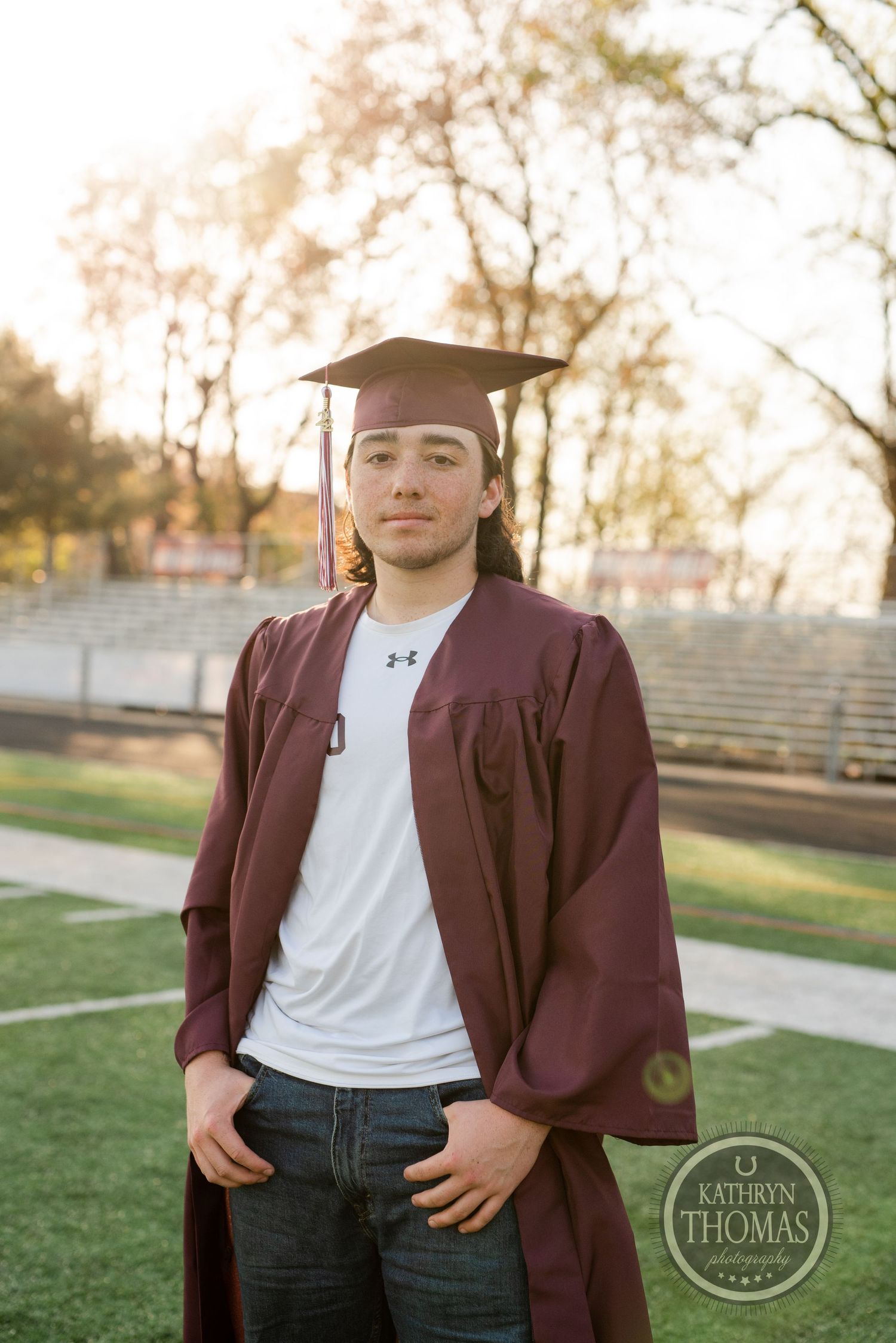 Graduate in maroon gown and mortarboard poses at sunset on football field for senior portraits.