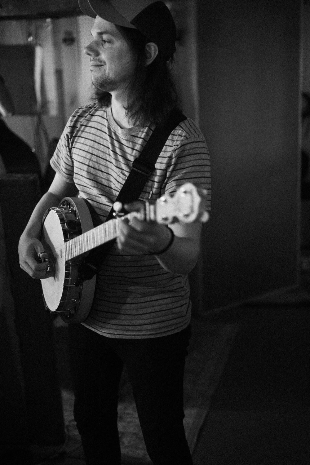Black and white photo of musician playing banjo while wearing a striped shirt and baseball cap.