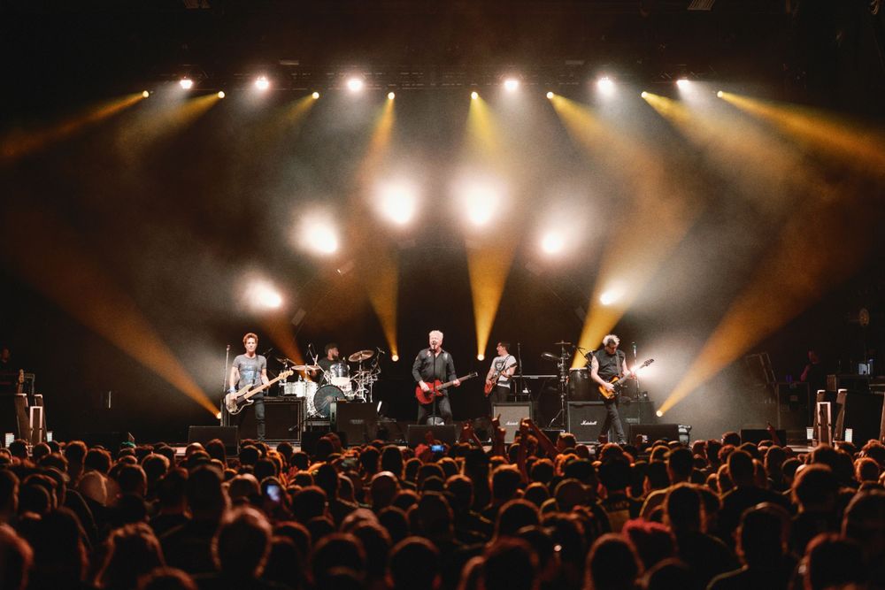 Concert stage with full band performing under dramatic orange and white spotlights.