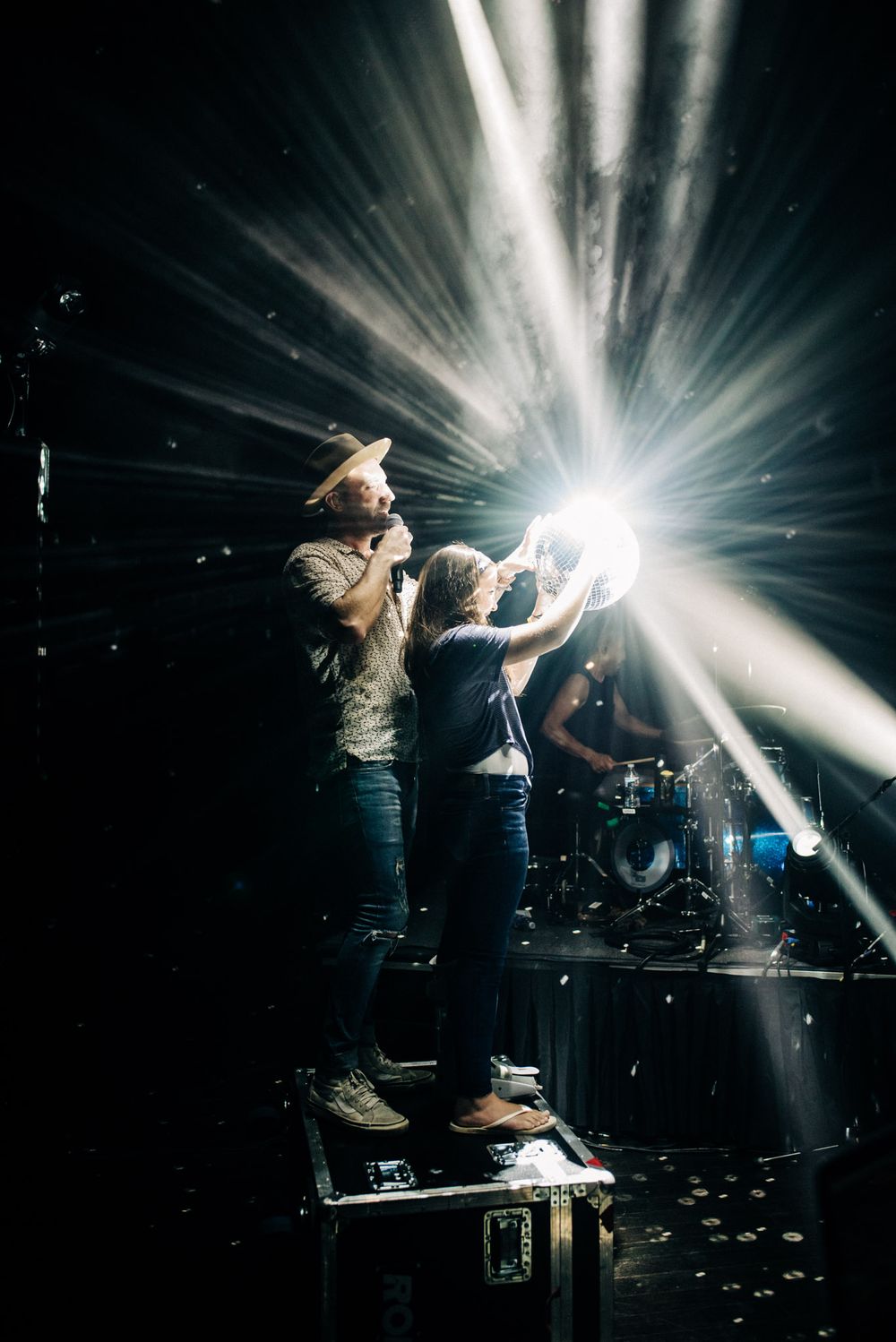 Performers stand elevated on platform with dramatic white light beams creating starburst effect.