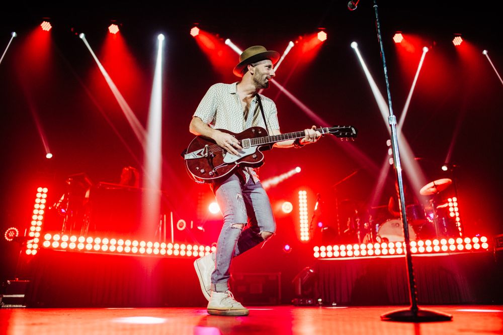 Energetic stage performance with guitarist in white outfit against red-lit backdrop.