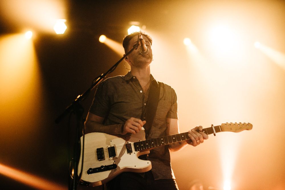 Musician plays white Telecaster guitar under golden stage lights.