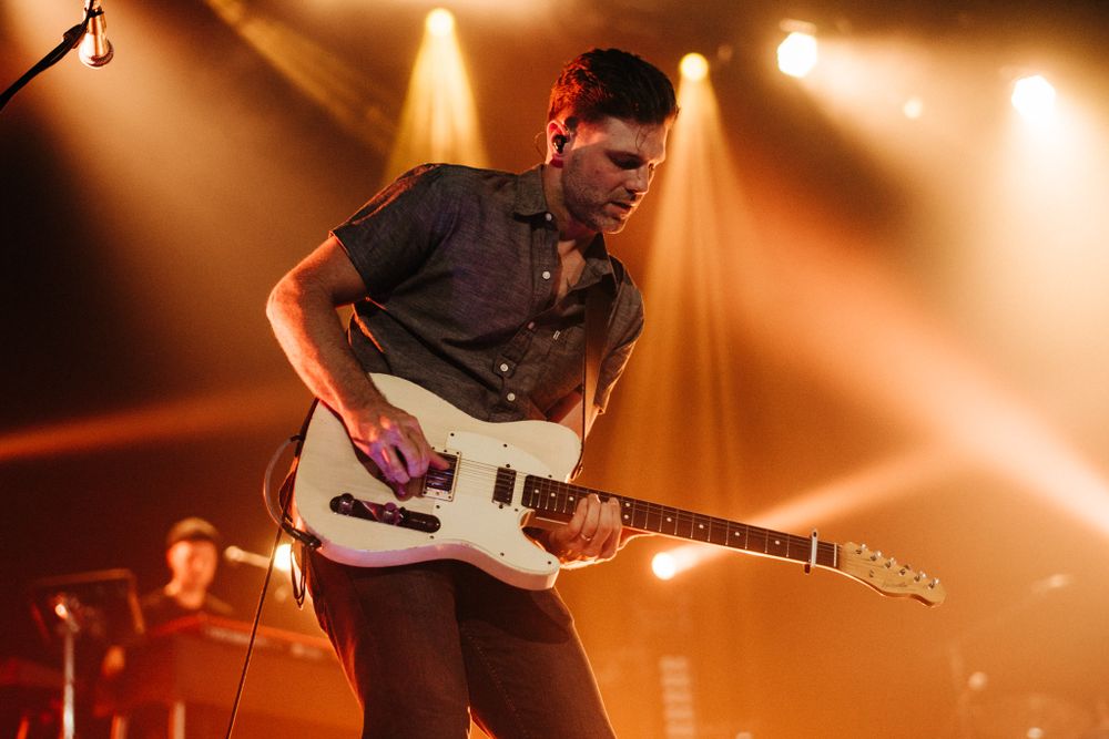Guitar player performs energetically with white electric guitar under orange lighting.