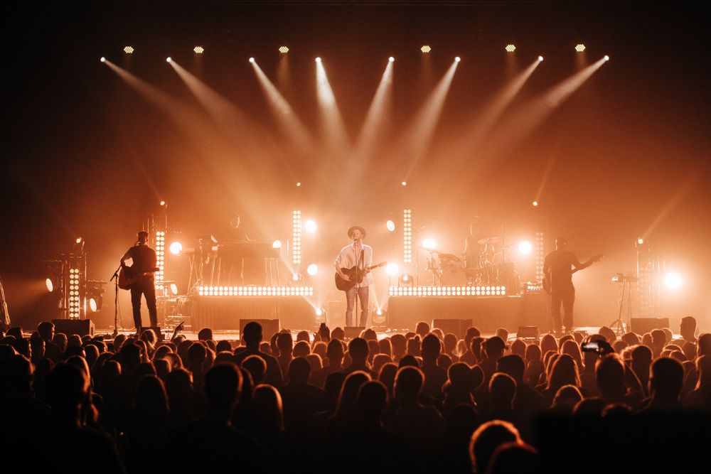 Musicians on stage with warm orange lighting and crowd silhouettes at live concert venue.