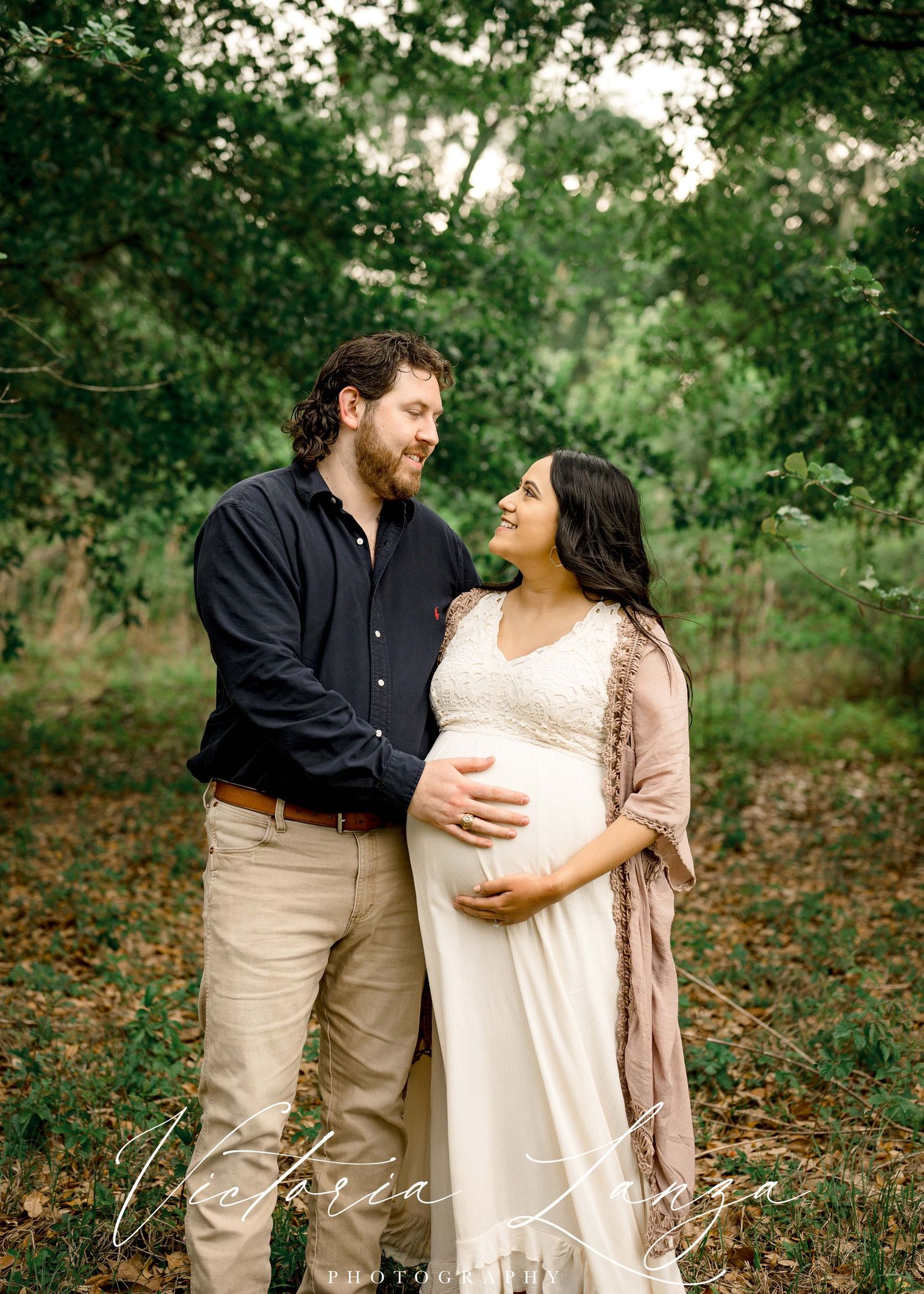 maternity portrait of pregnant wife looking at husband while embracing belly in a greenery outdoor space
