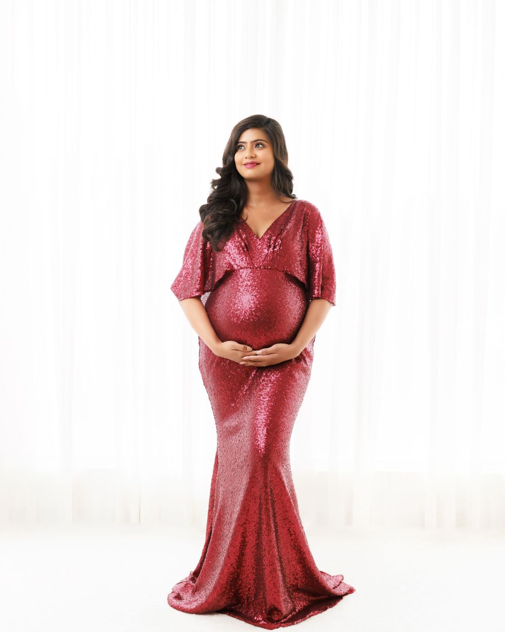 Elegant maternity portrait in form-fitting red sequin gown against white background.