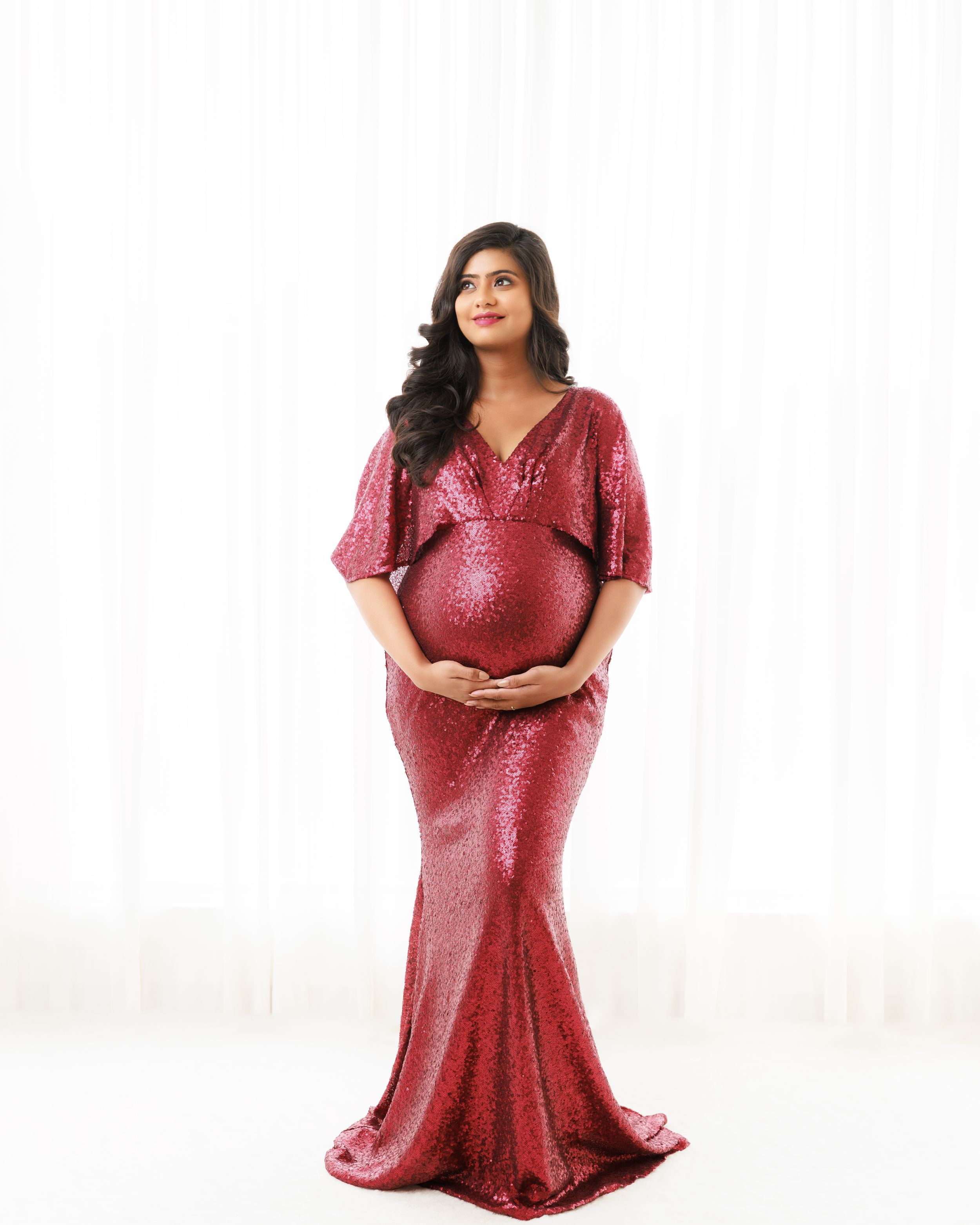 Elegant maternity portrait in form-fitting red sequin gown against white background.