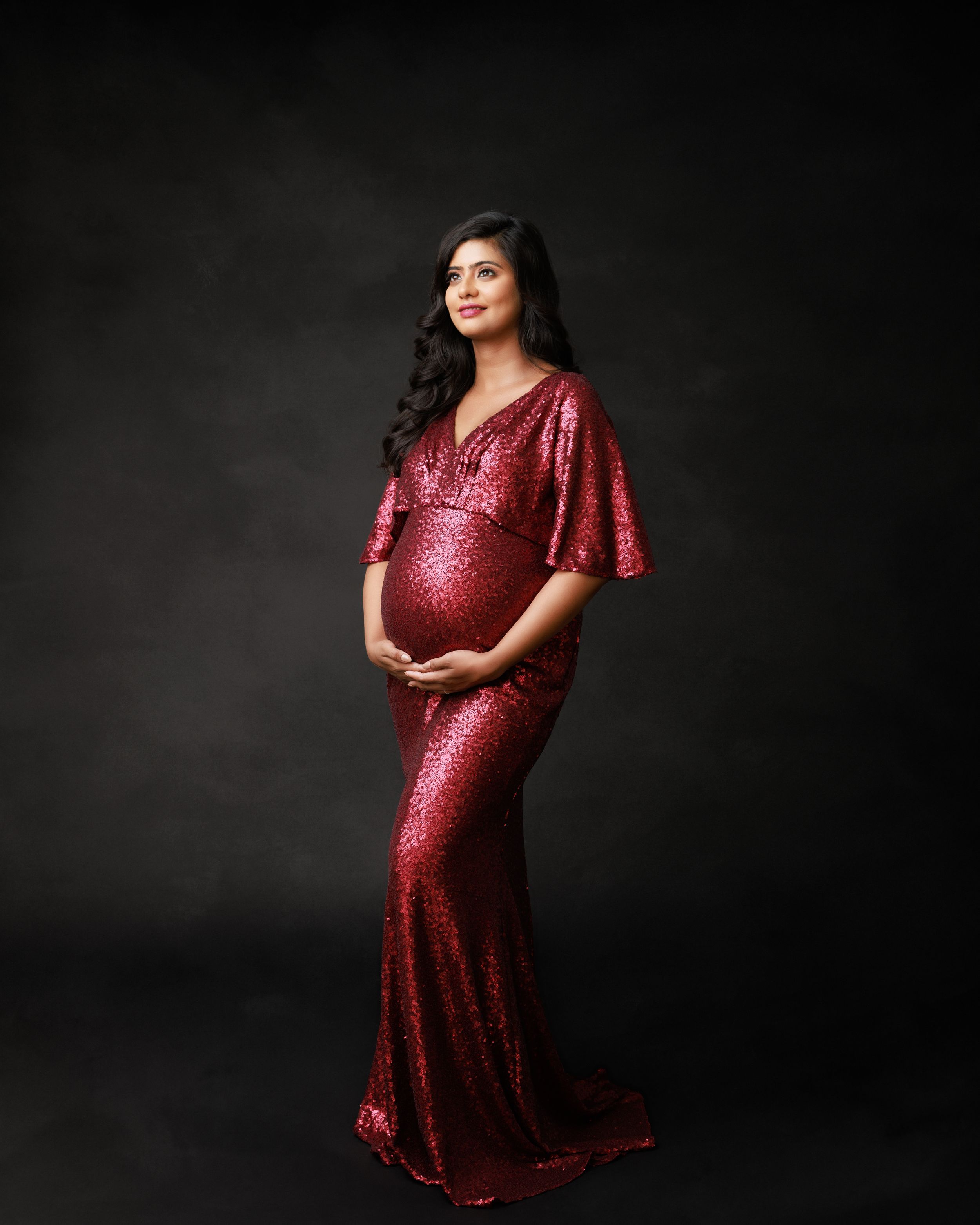Dramatic maternity portrait in sparkly red gown against dark background.