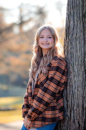 A portrait of someone wearing a plaid flannel shirt leaning against a tree trunk in autumn.