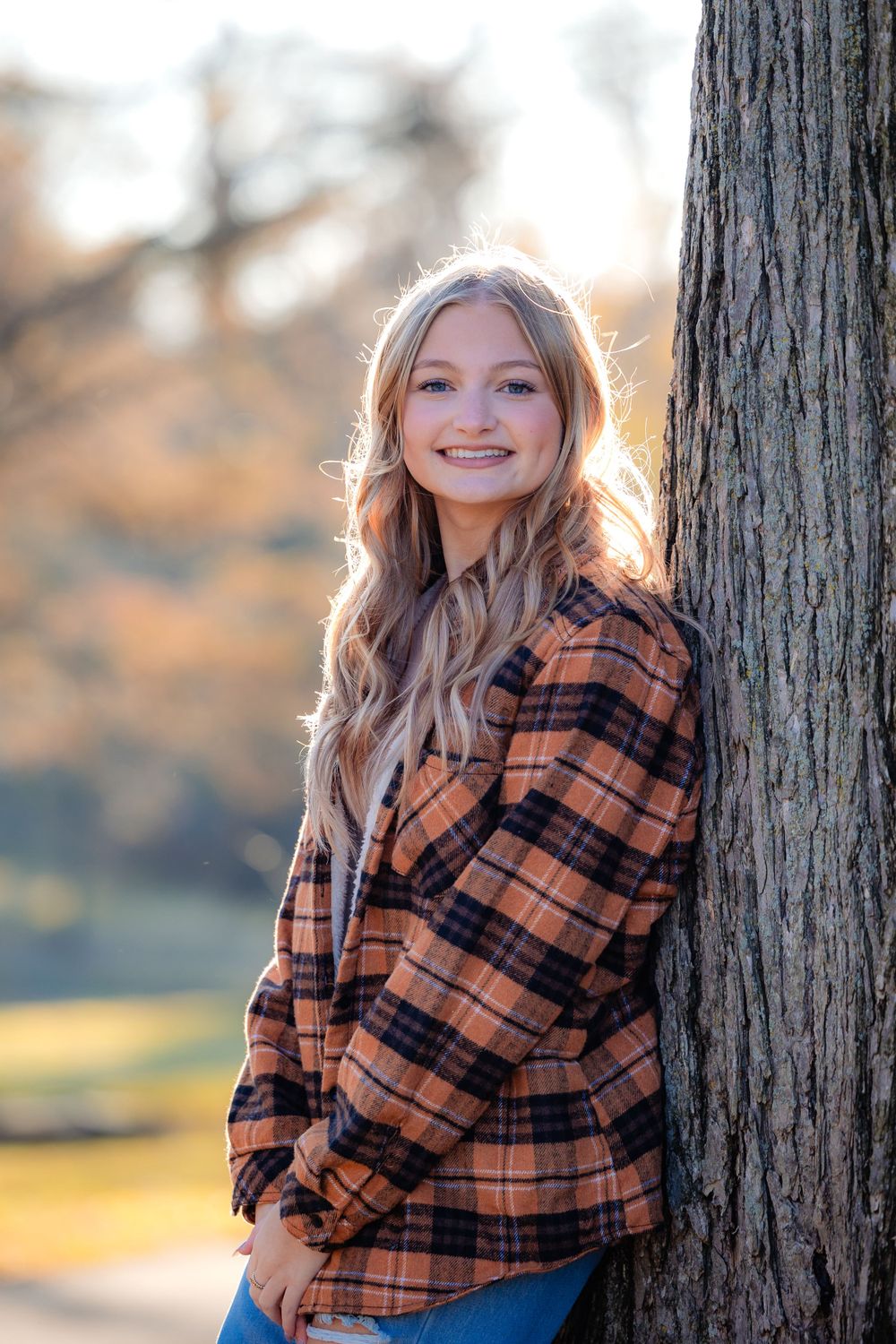 A portrait of someone wearing a plaid flannel shirt leaning against a tree trunk in autumn.