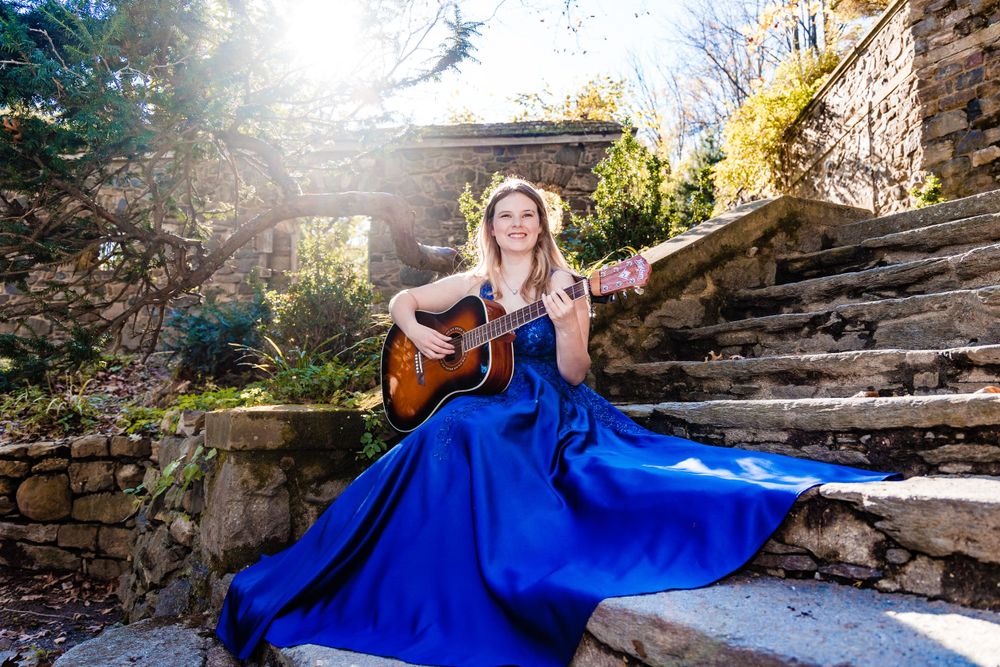 Musician in blue formal dress plays acoustic guitar while seated on stone steps in sunlit garden setting.