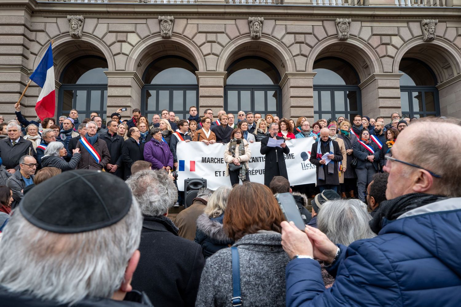 Crowd gathered for a protest demonstration outside a historic building with arched architecture.