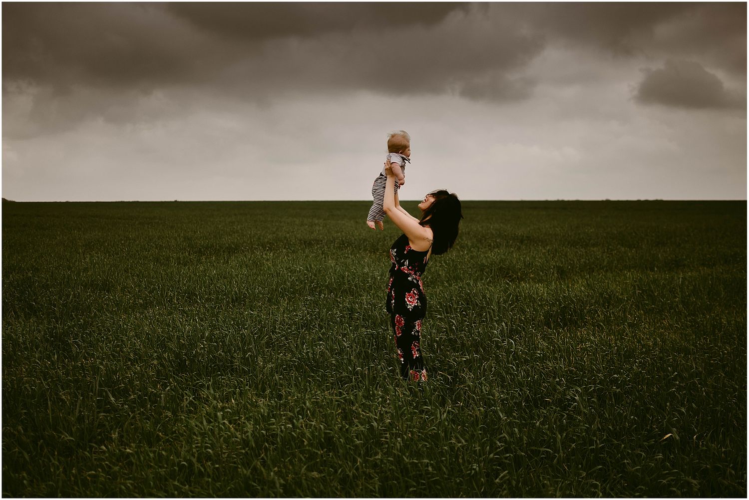 Silhouette of parent lifting baby joyfully against moody sky in open field.