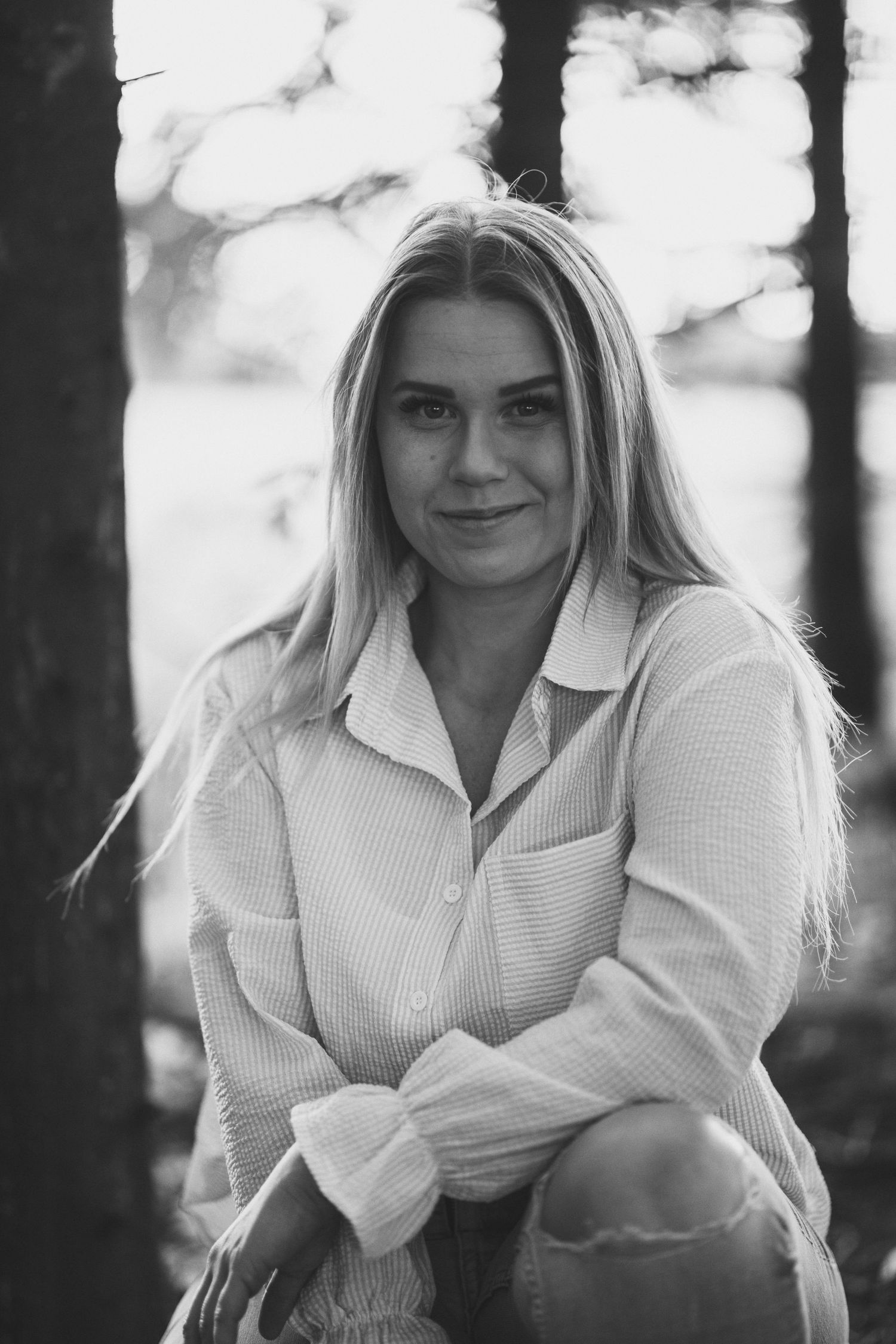 Black and white portrait of a woman in a white button-down shirt among pine trees.