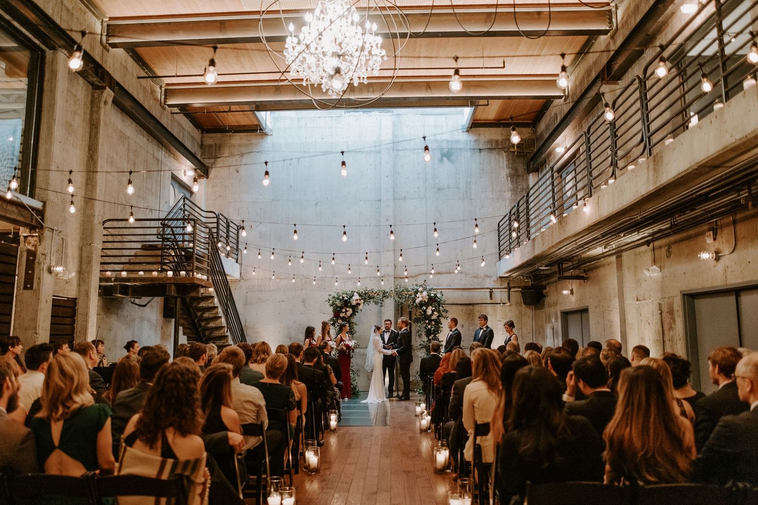 Modern industrial wedding ceremony venue with hanging chandelier and gathered guests watching the celebration.