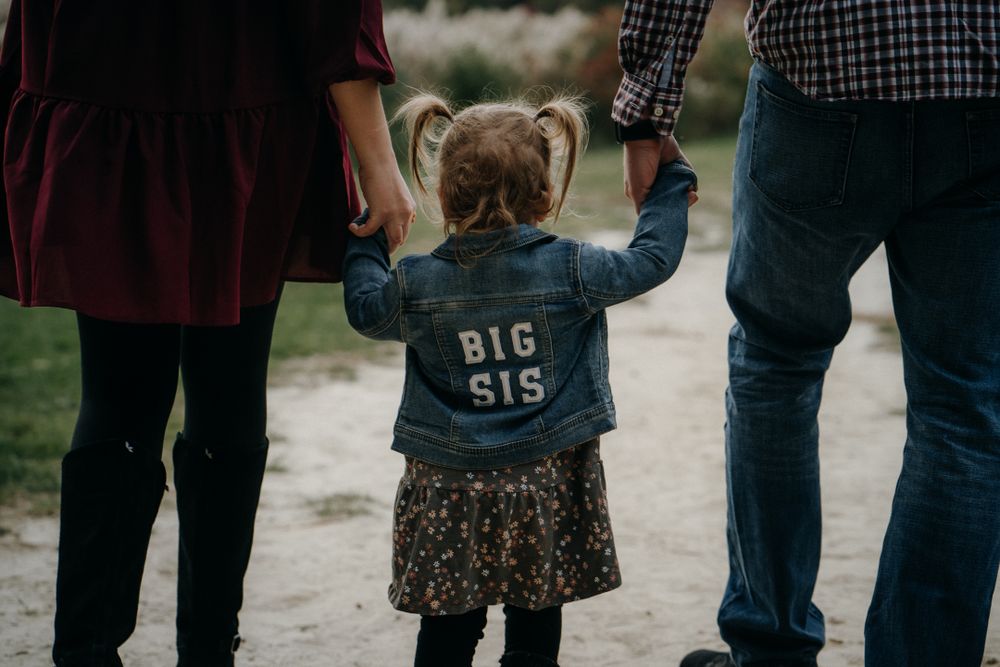 Child wearing 'Big Sis' jacket holding parents' hands outdoors.