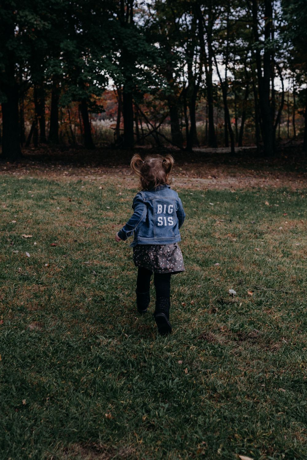 Child in 'Big Sis' denim jacket standing in wooded area at sunset.