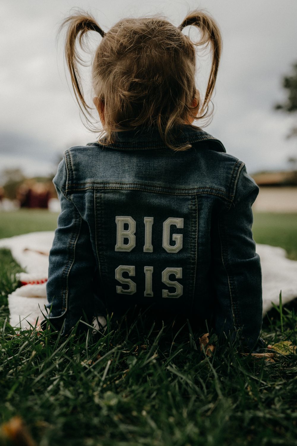 Back view of child wearing denim jacket with 'BIG SIS' text sitting on grass.