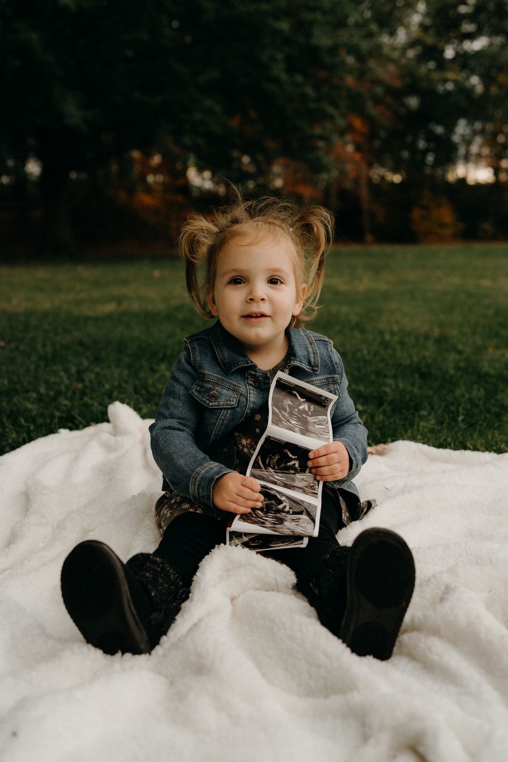 Small child in denim jacket sitting on white blanket holding ultrasound photos.