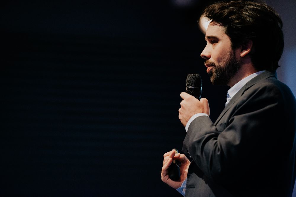 A speaker in a dark suit presenting on stage with microphone against black background.