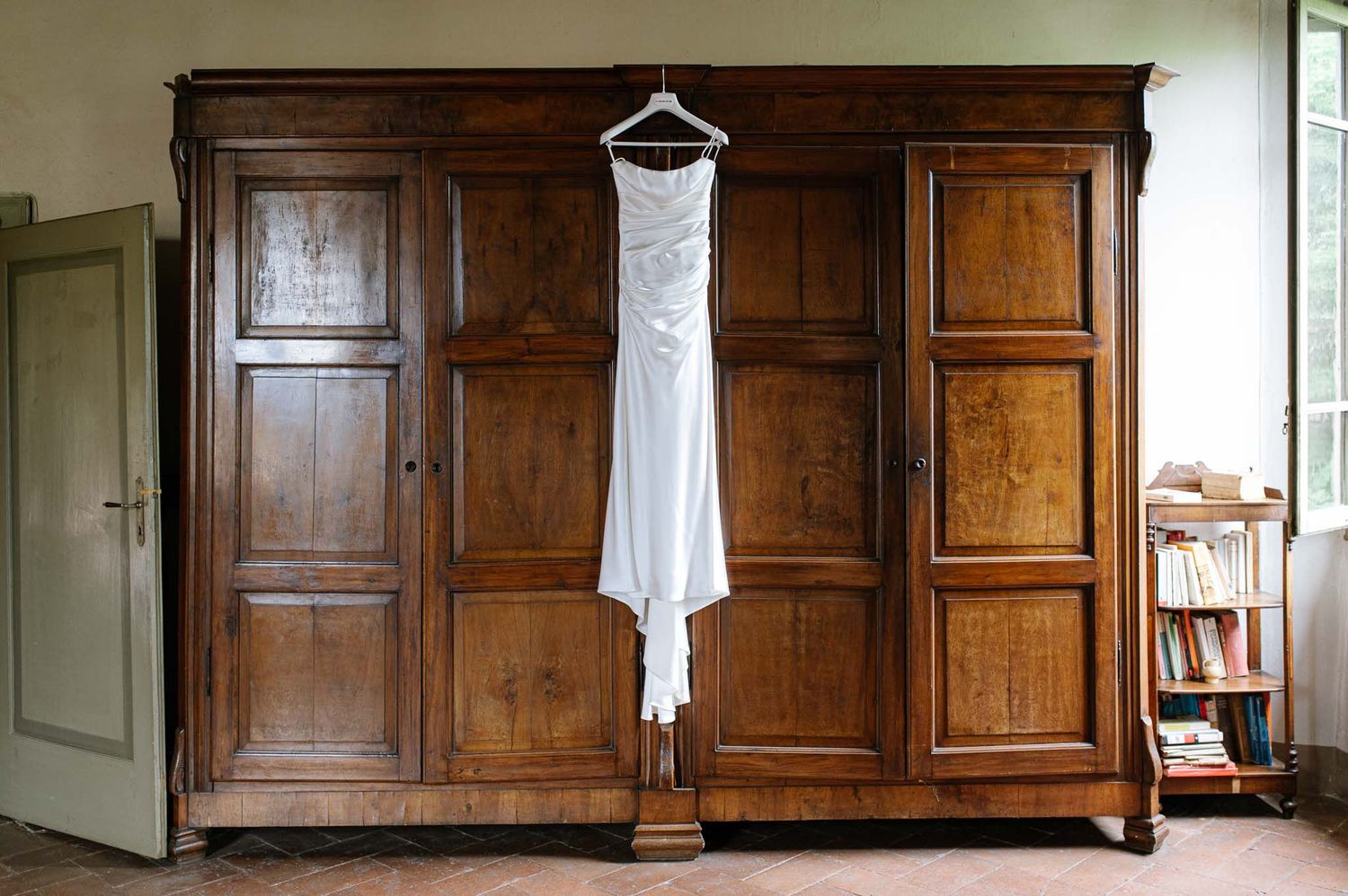 White wedding dress hanging on wooden antique wardrobe in natural light.