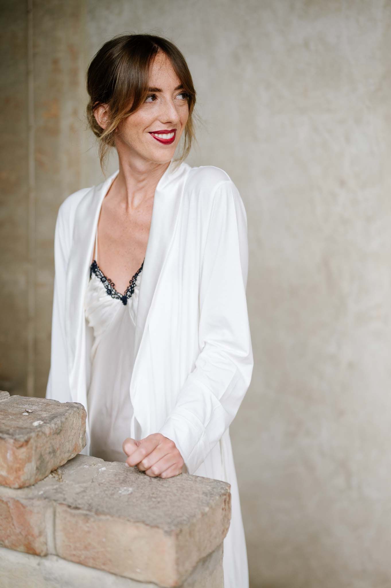 A figure in white clothing smiles while leaning on a stone wall.