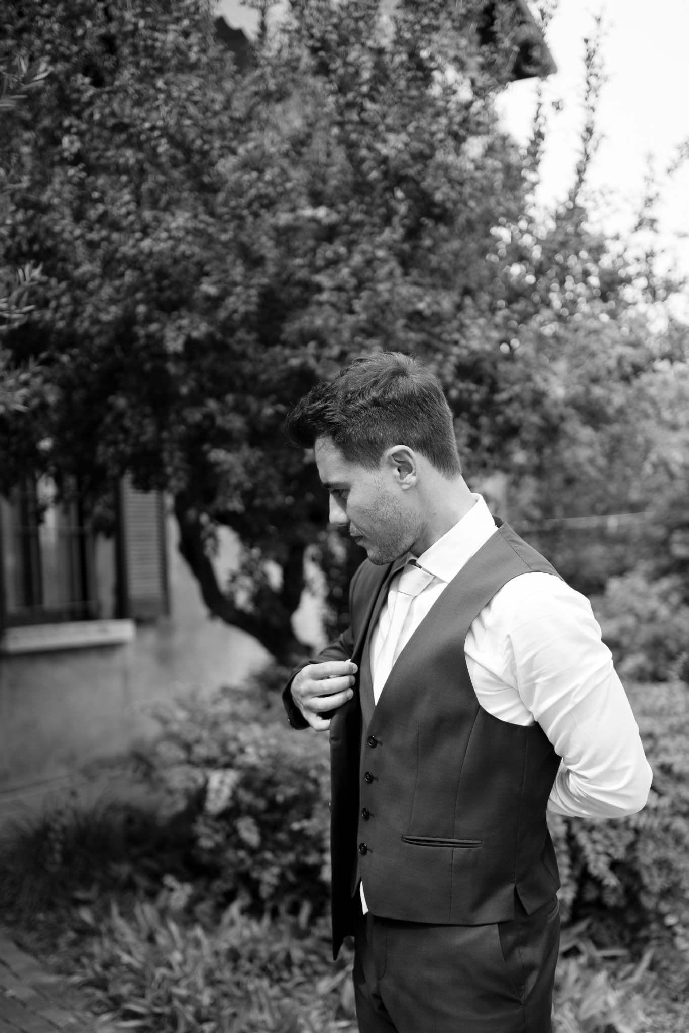 A black and white portrait of someone adjusting formal attire in a garden setting.