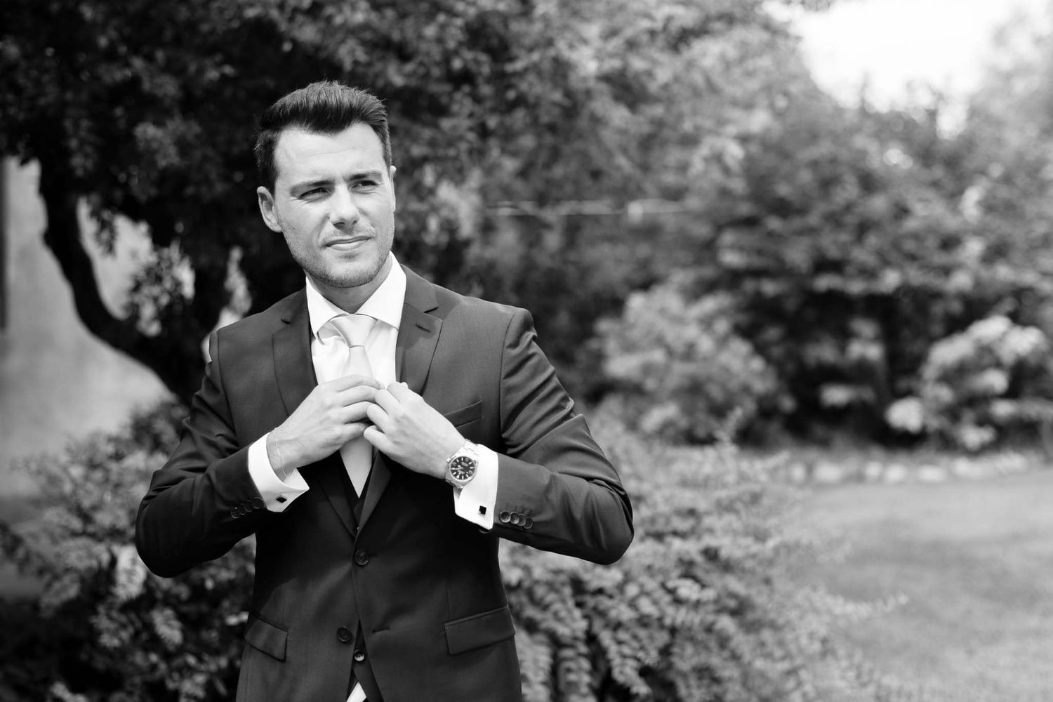 A black and white portrait showing someone adjusting a formal tie outdoors.