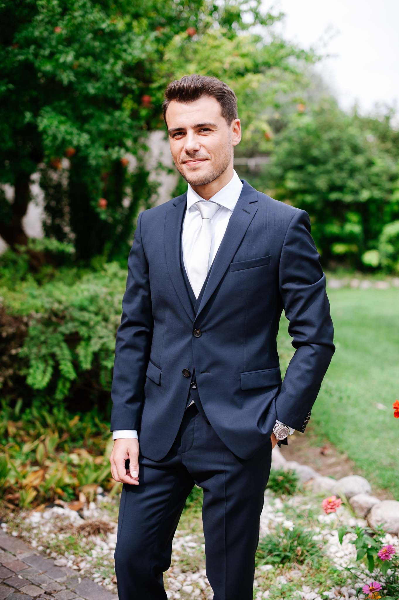 A portrait of someone in a tailored navy suit standing in a lush garden setting.