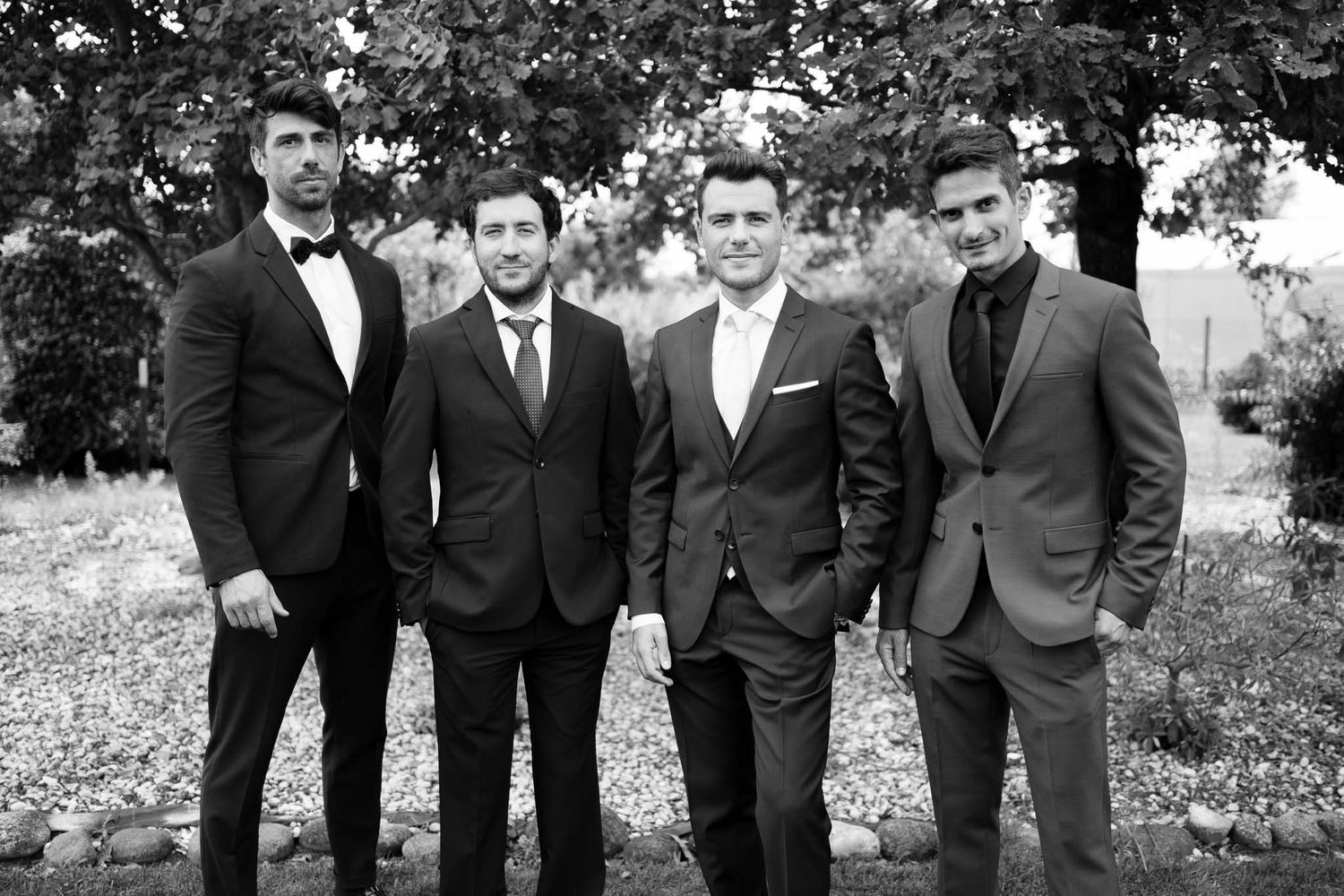 Four groomsmen in formal suits pose together outdoors in black and white photography.