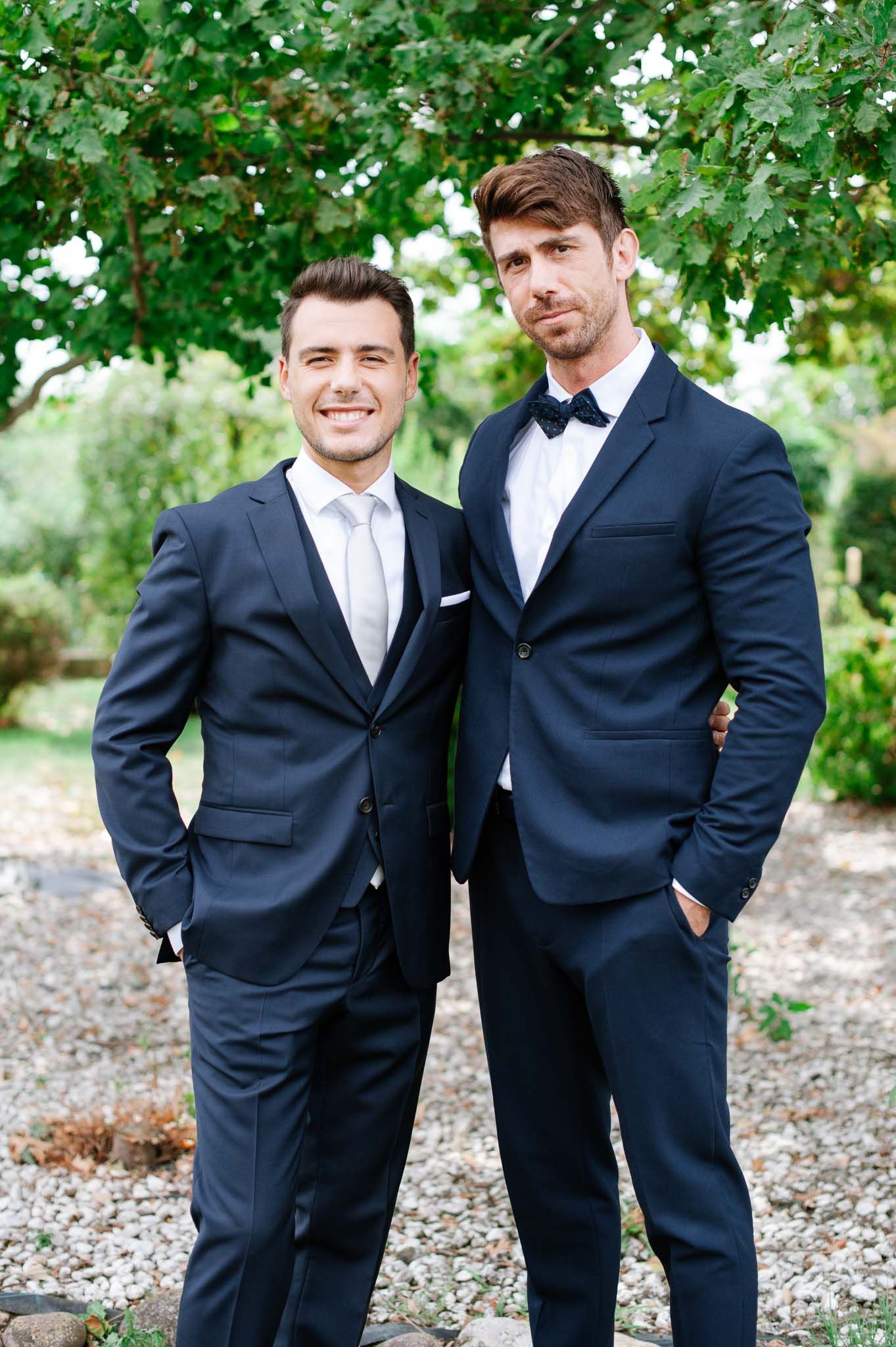 Two gentlemen in navy suits and bow ties pose together in garden setting.
