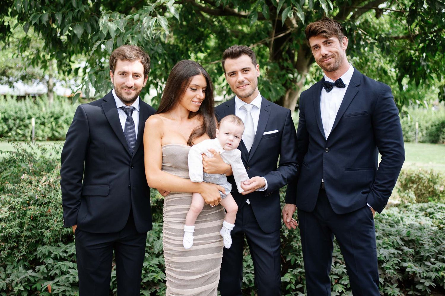 Group portrait of groomsmen in formal attire with a baby at an outdoor wedding.