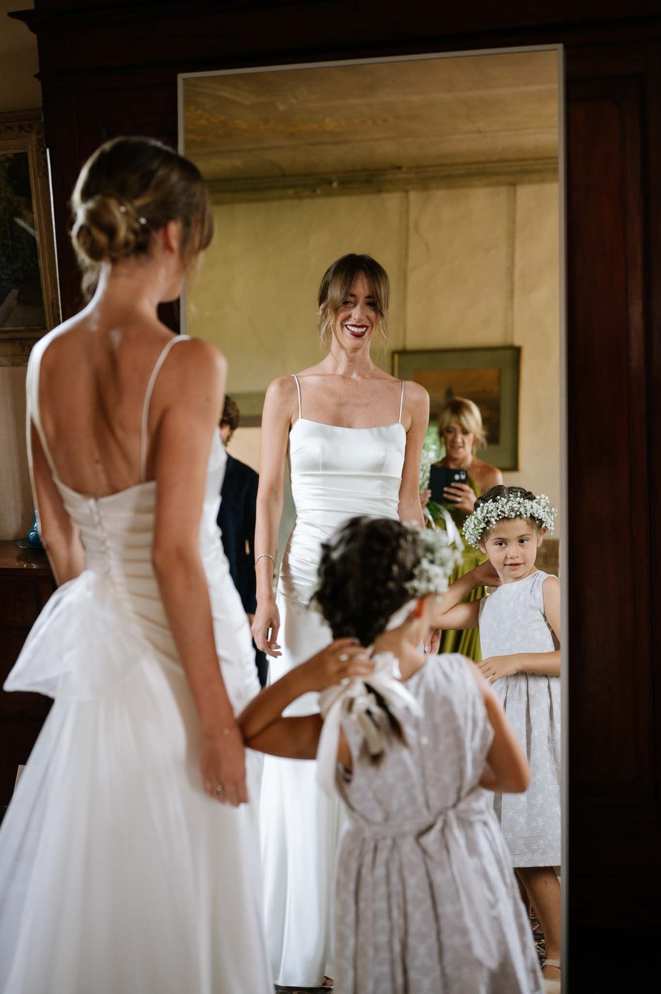Bride in white dress looks in mirror while flower girl watches during wedding preparations.