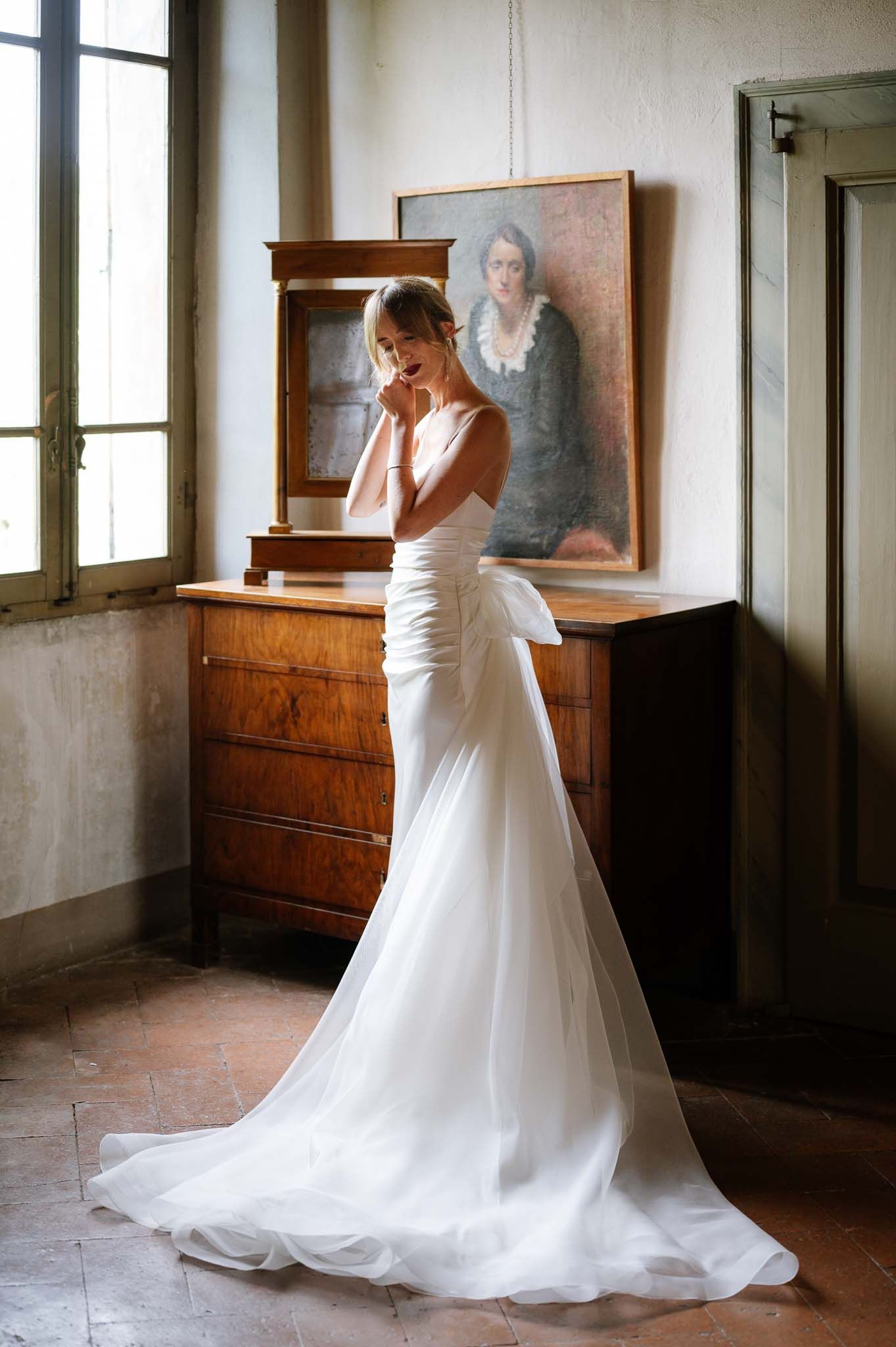 Elegant bride in fitted white gown poses beside vintage wooden dresser with mirror.