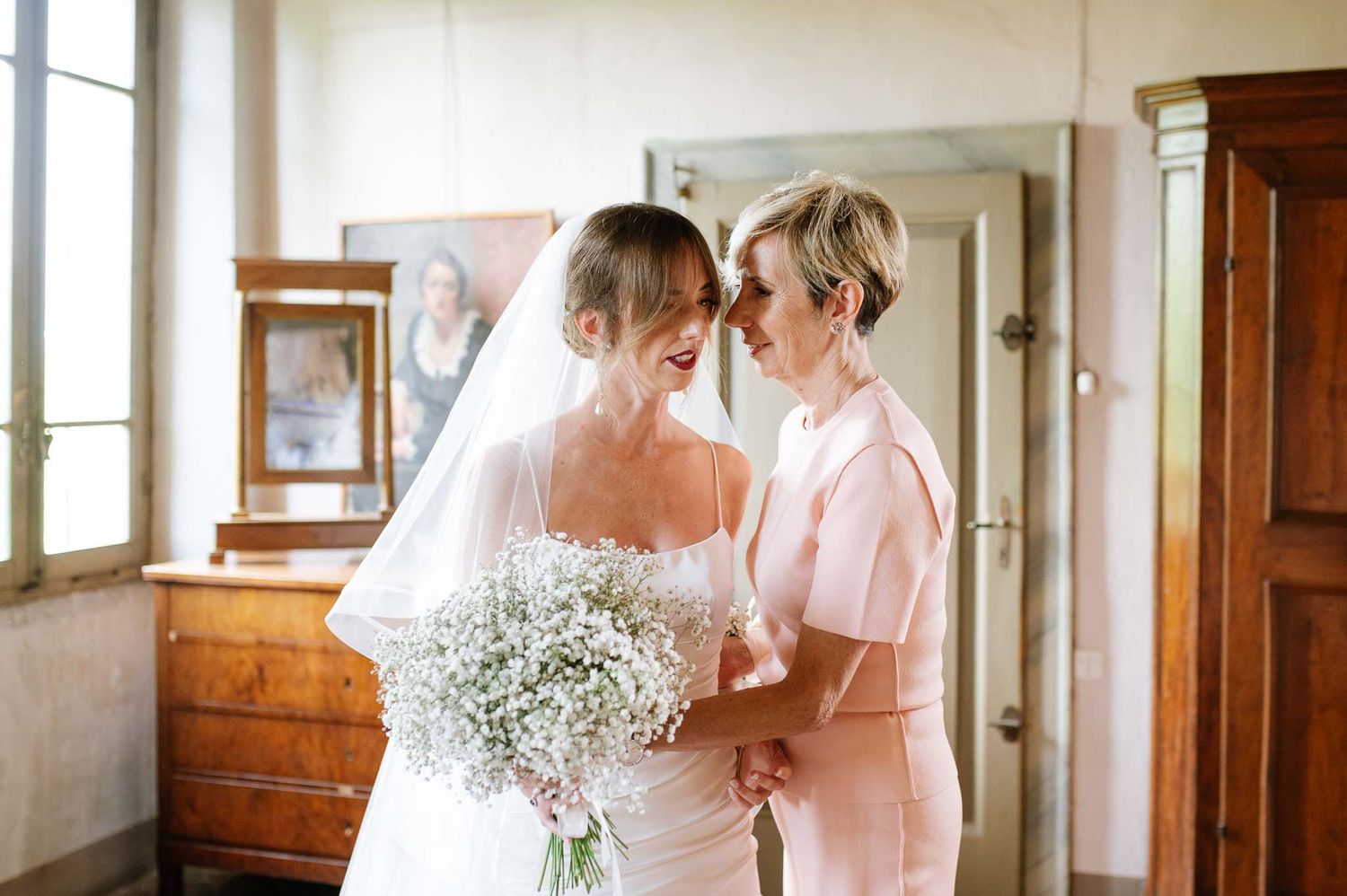 Tender moment between bride in white gown and companion holding baby's breath bouquet.
