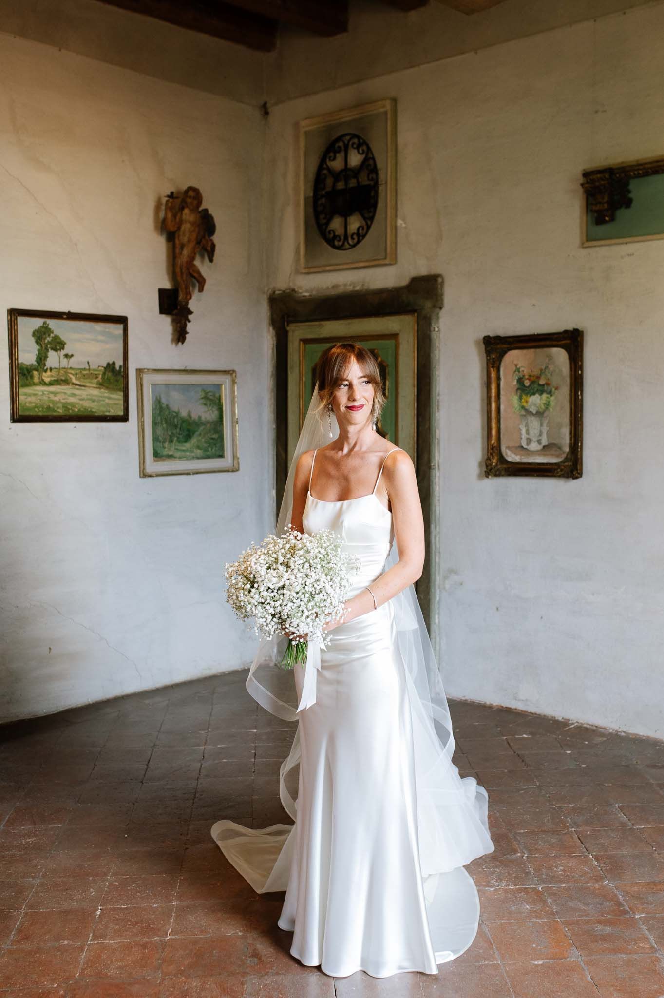 Elegant bride in sleek white gown poses with baby's breath bouquet against rustic wall with vintage artwork.