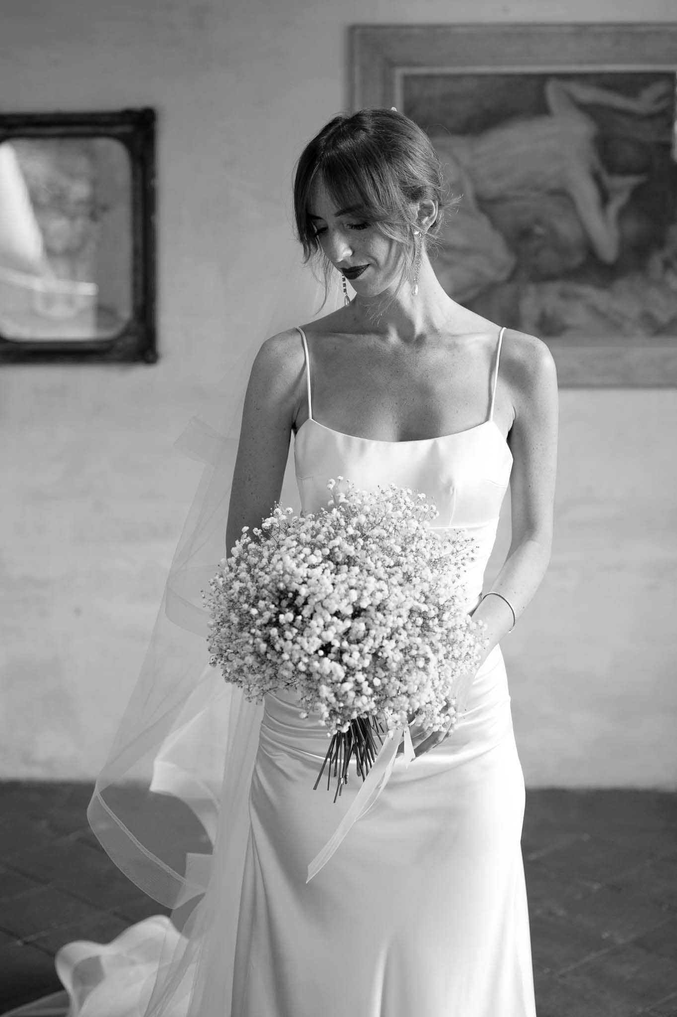 Black and white photo of bride in simple white gown holding baby's breath bouquet in historic venue.