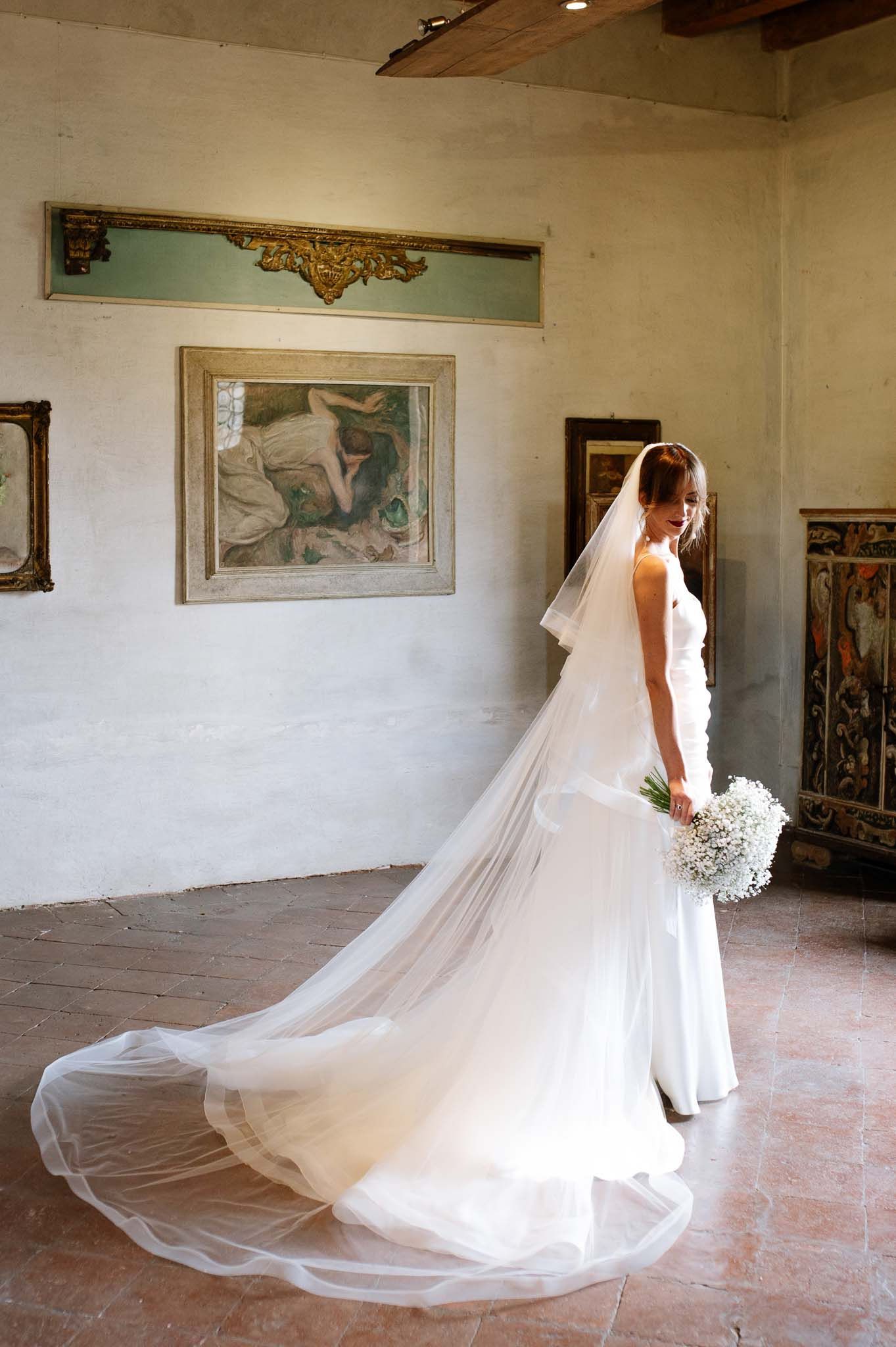 Bride in elegant white gown with flowing veil poses against rustic wall with vintage artwork.