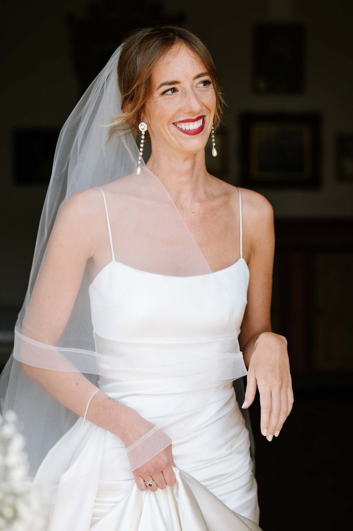 Bride in white spaghetti strap gown and veil smiles joyfully during wedding preparations.