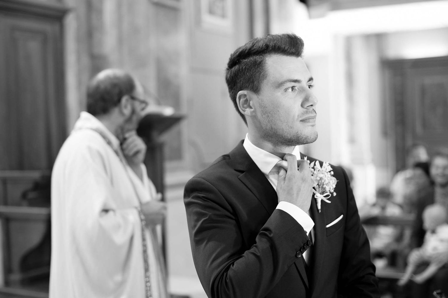 A gentleman in formal attire adjusts his bowtie in a black and white church setting.